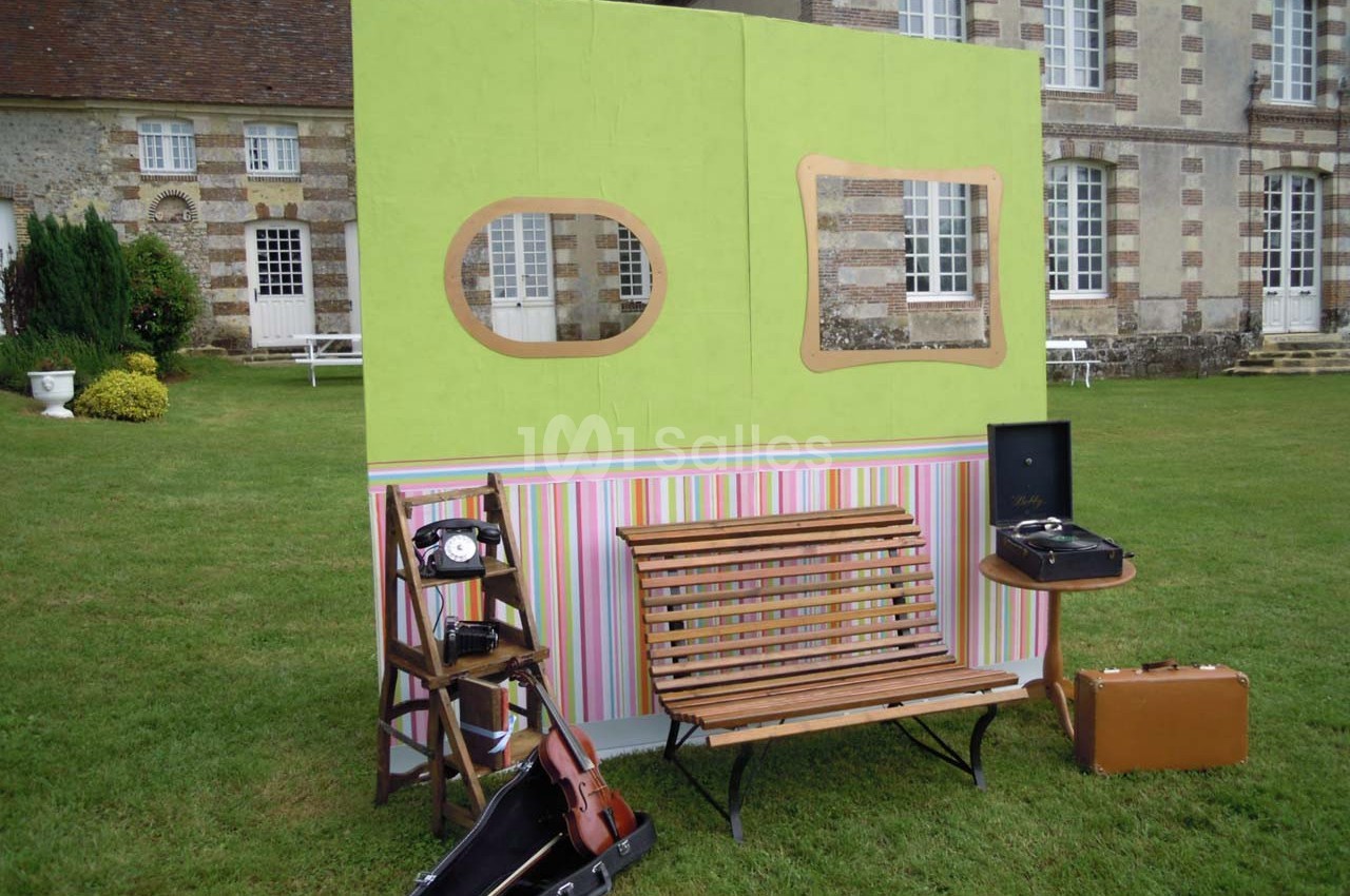 Décor vintage en extérieur avec banc, violon, téléphone ancien, valise, tourne-disque et façade colorée devant un bâtiment.