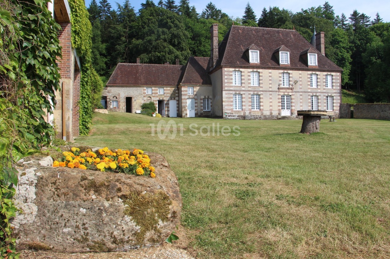 Manoir en pierre entouré de pelouse, avec un massif de fleurs jaunes au premier plan et une forêt en arrière-plan.
