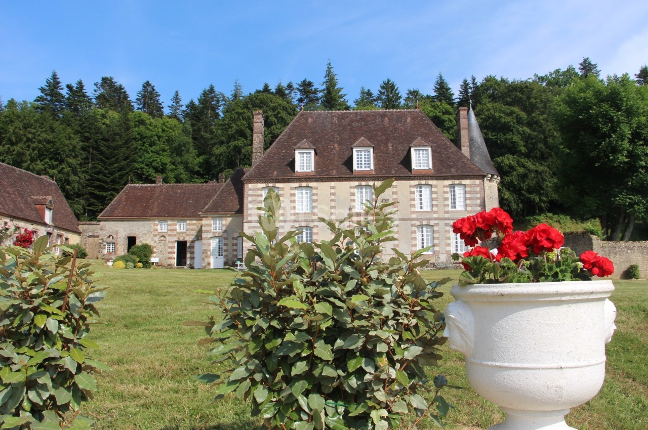 Manoir en pierre entouré de verdure, avec des rosiers et une jardinière blanche au premier plan.
