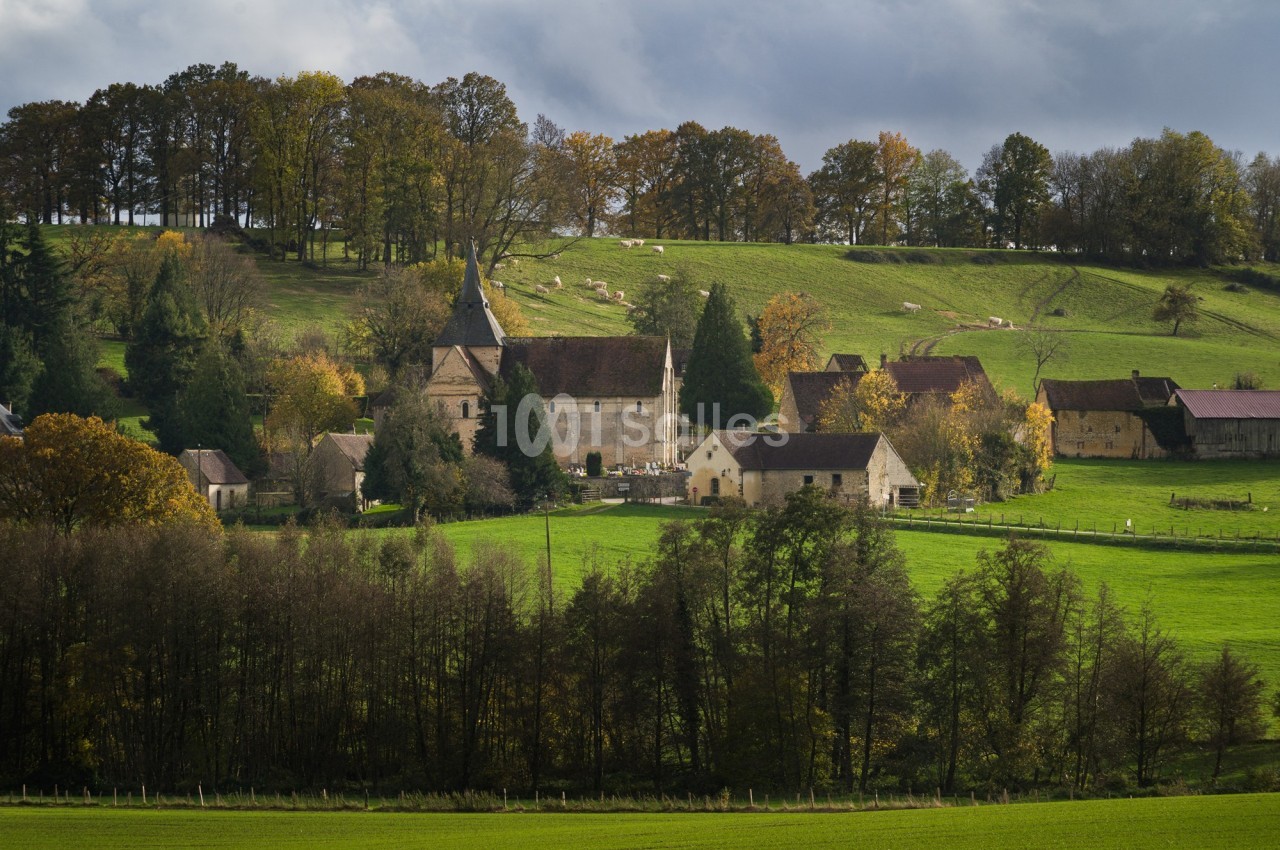 Village rural avec église en pierre entourée de prairies verdoyantes, arbres et bâtiments traditionnels sous un ciel nuageux.