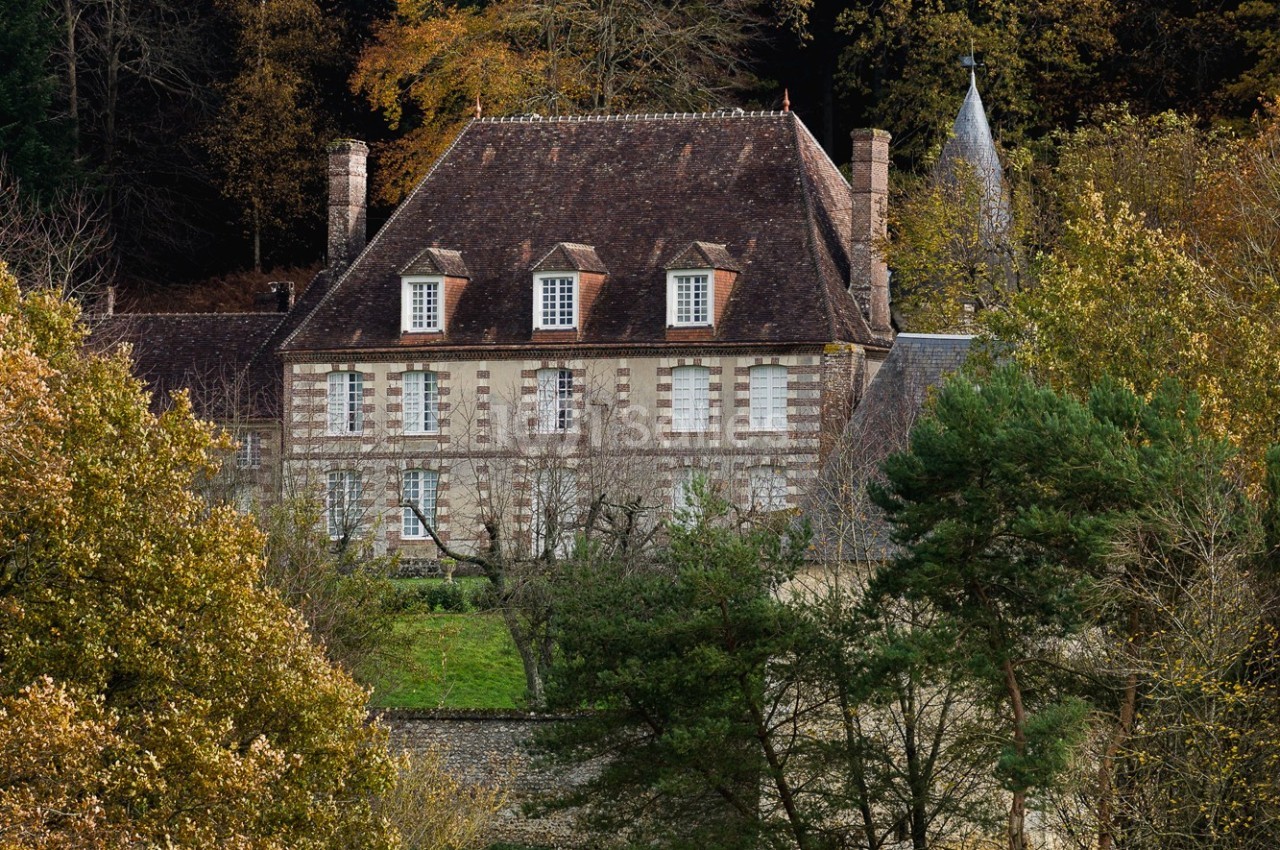 Manoir en pierre entouré d'arbres et de végétation, avec un toit en tuiles et des fenêtres à petits carreaux.