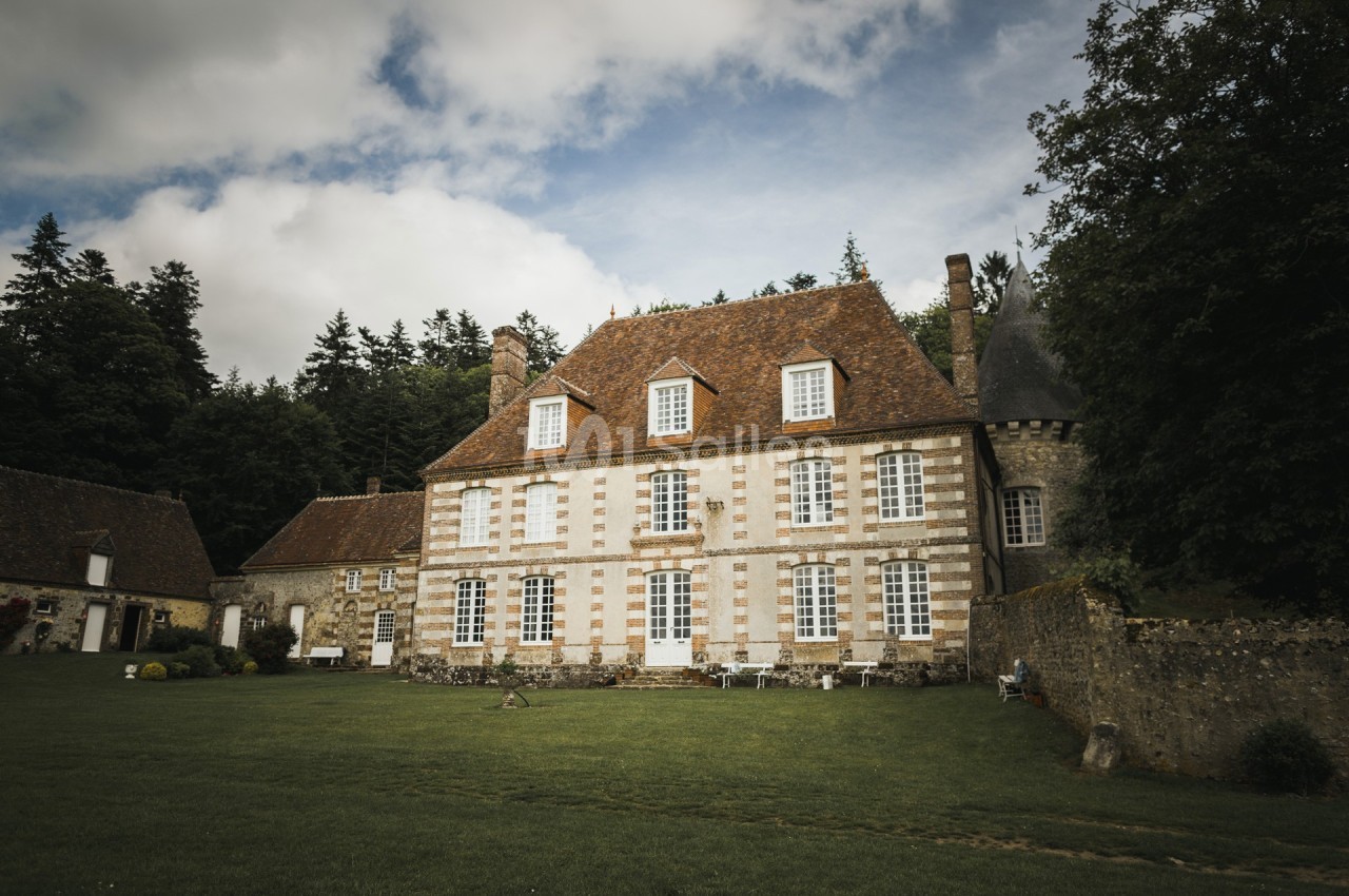 Grande maison en pierre avec toit en tuiles, entourée de pelouse et d'arbres, sous un ciel partiellement nuageux.