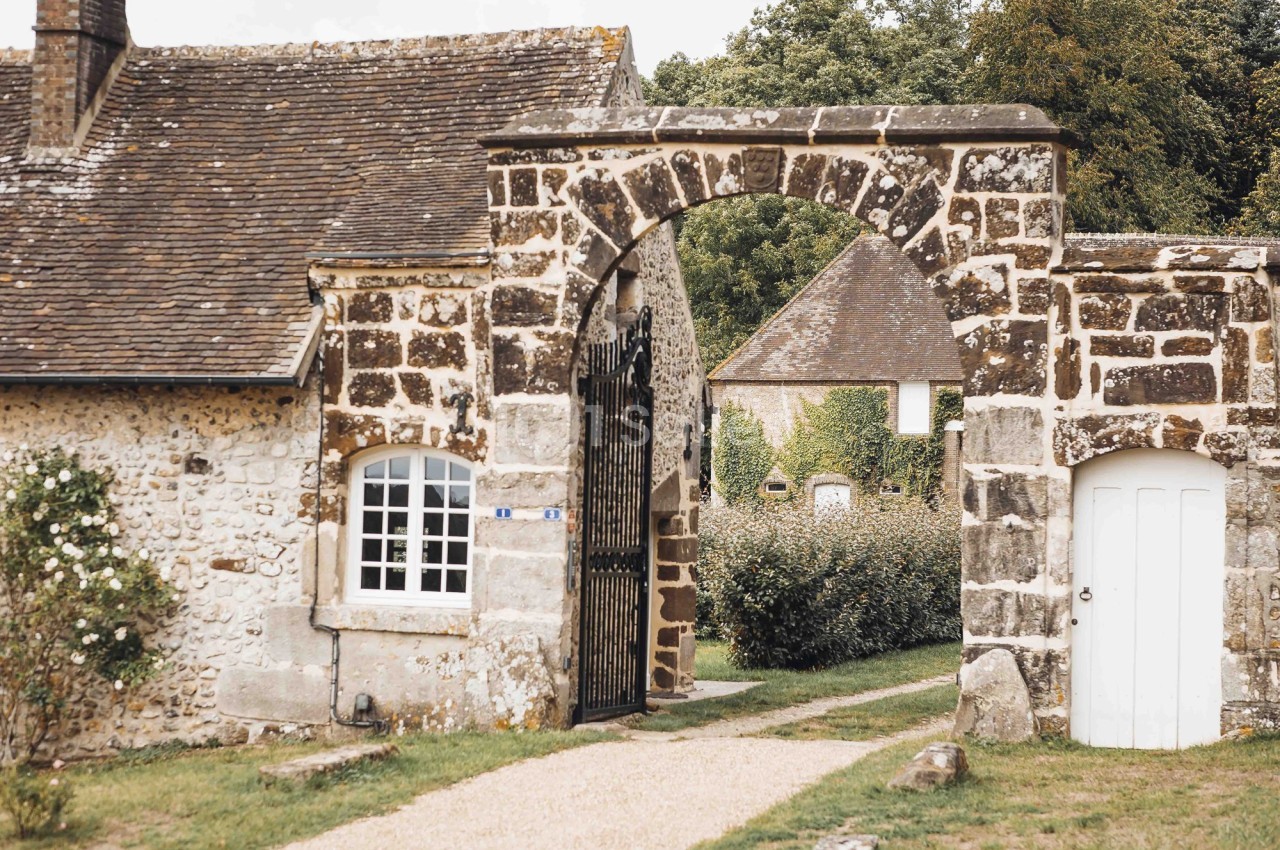 Portail en pierre avec arche, menant à une maison en pierre et un jardin verdoyant en arrière-plan.