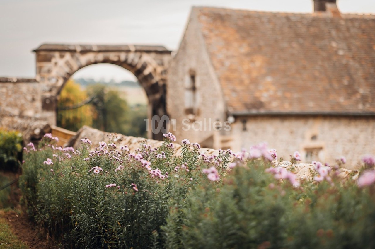 Fleurs violettes en premier plan devant un bâtiment en pierre et une arche dans un paysage rural.
