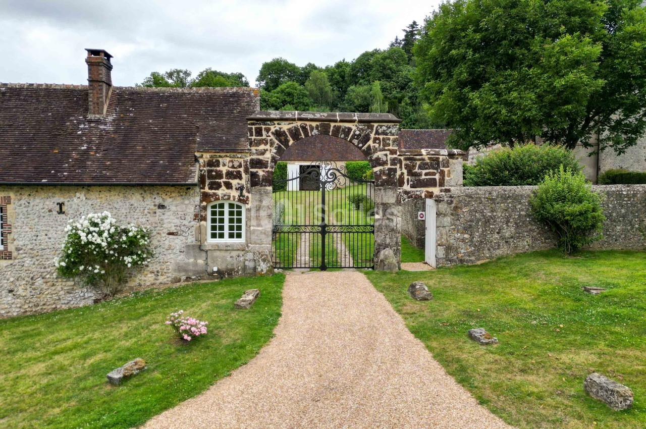Portail en fer forgé encadré par des bâtiments en pierre, donnant sur une allée gravillonnée et un jardin verdoyant.