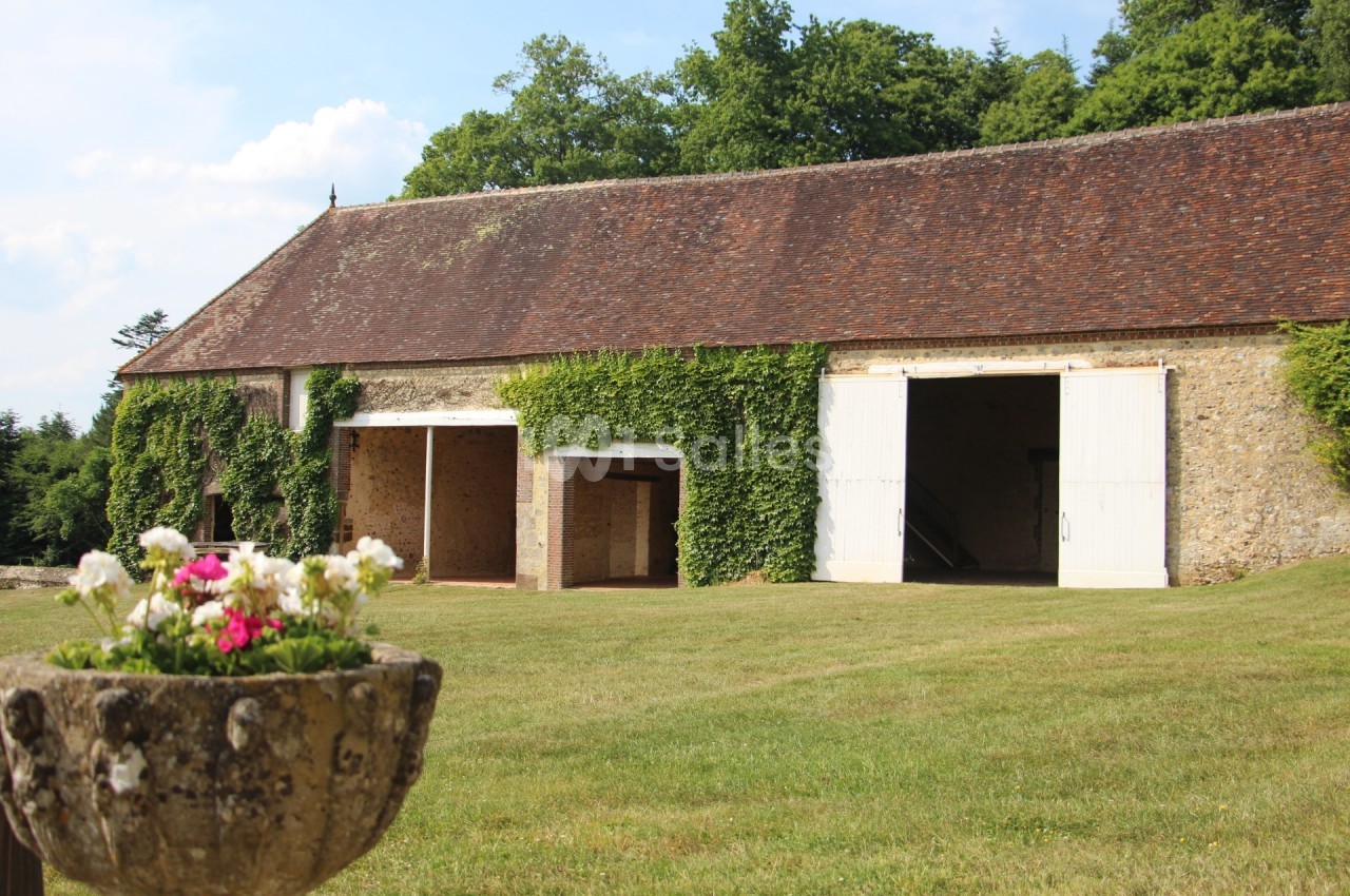 Grange en pierre avec toit en tuiles, portes blanches ouvertes et murs partiellement couverts de lierre, entourée de pelouse.