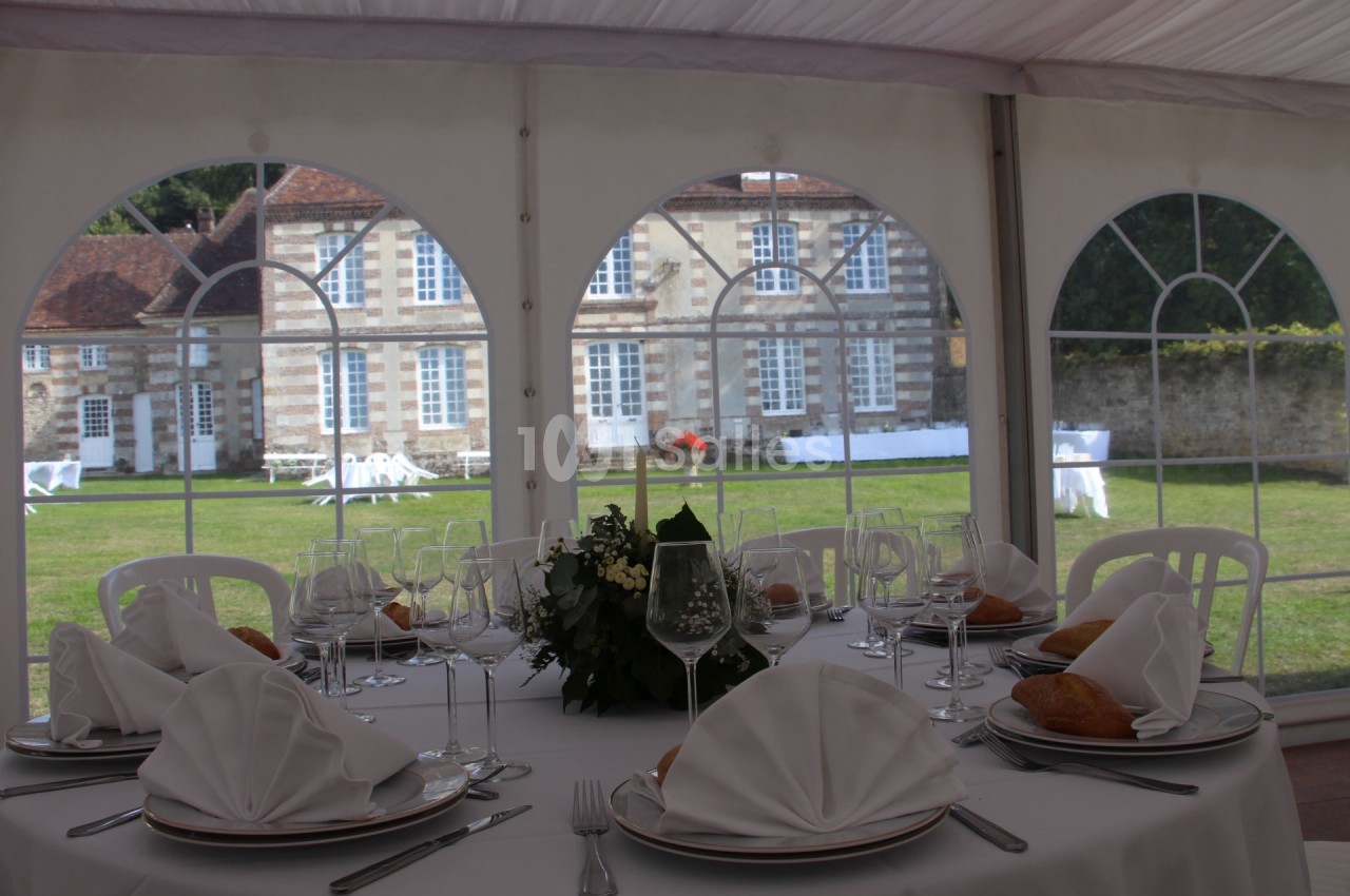Table dressée avec nappes blanches, serviettes pliées et verres, sous une tente avec vue sur un bâtiment ancien et un jardin.