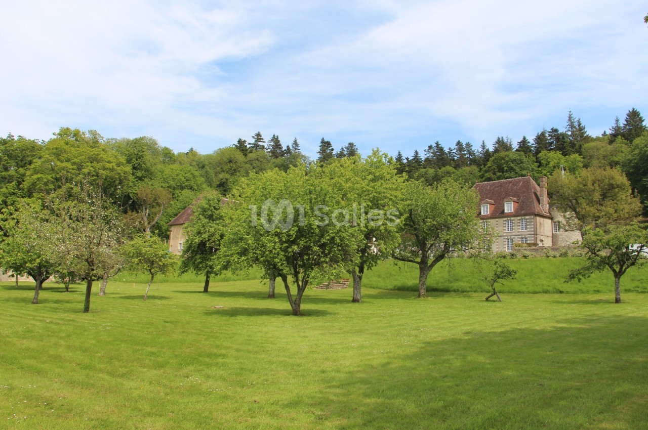 Grand jardin verdoyant avec des arbres dispersés, une maison en pierre et une forêt en arrière-plan sous un ciel bleu.
