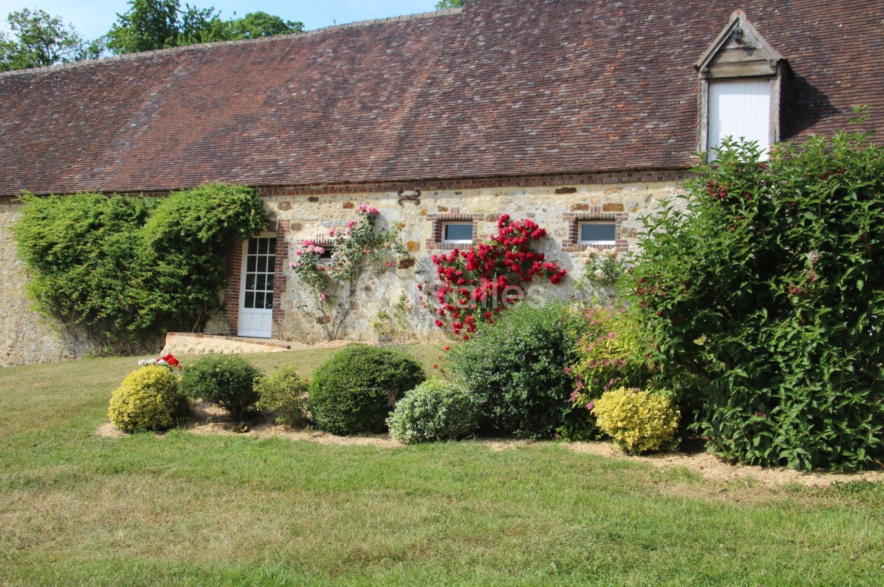 Façade en pierre d'une maison ancienne avec toit en tuiles, rosiers fleuris et jardin arboré au premier plan.
