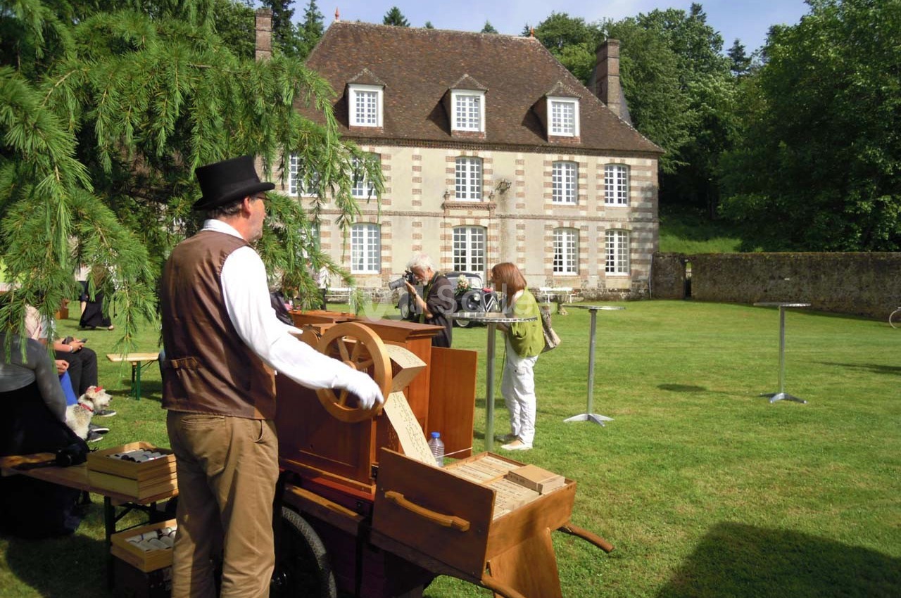 Un homme joue de l'orgue de barbarie dans un jardin devant une grande maison ancienne en pierre.