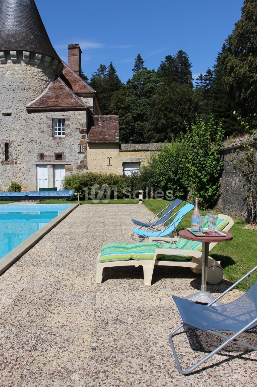 Chaises longues et table basse près d'une piscine, avec un bâtiment en pierre et des arbres en arrière-plan.