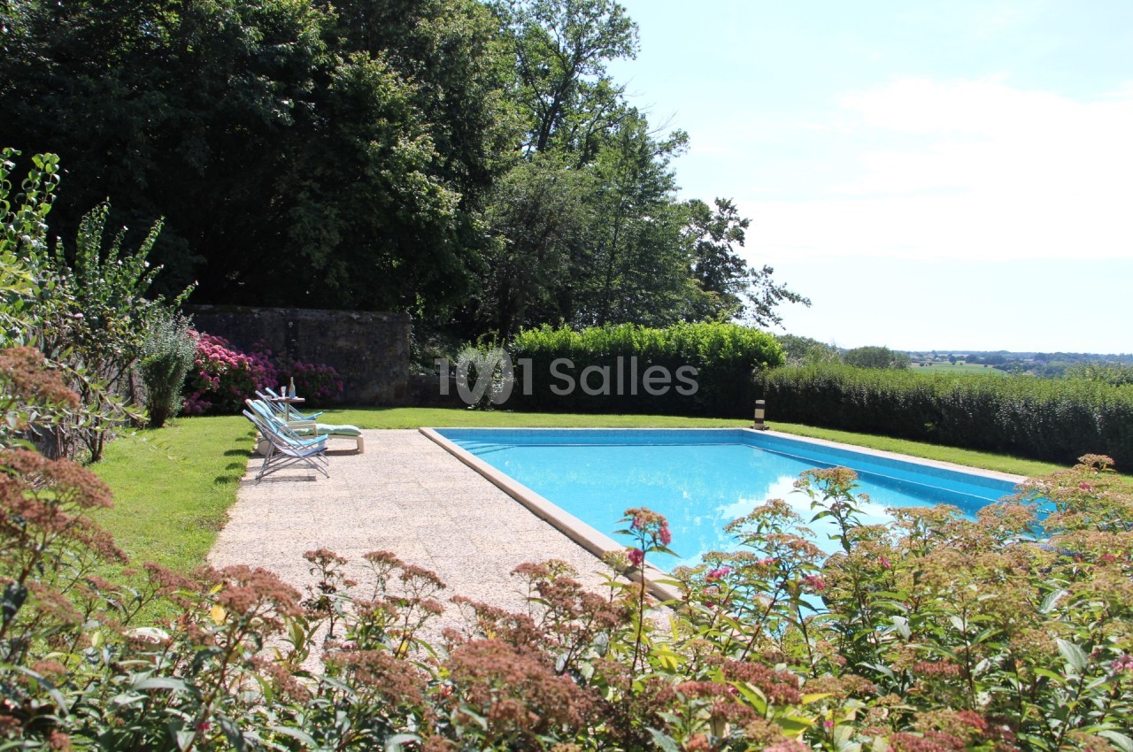Piscine extérieure entourée de verdure et de fleurs, avec des chaises longues sur une terrasse ensoleillée.