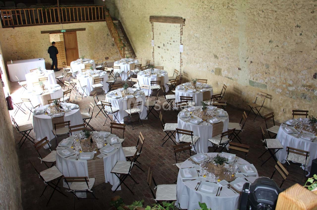 Salle de réception rustique avec des tables rondes dressées, nappes blanches et chaises en bois pliantes.