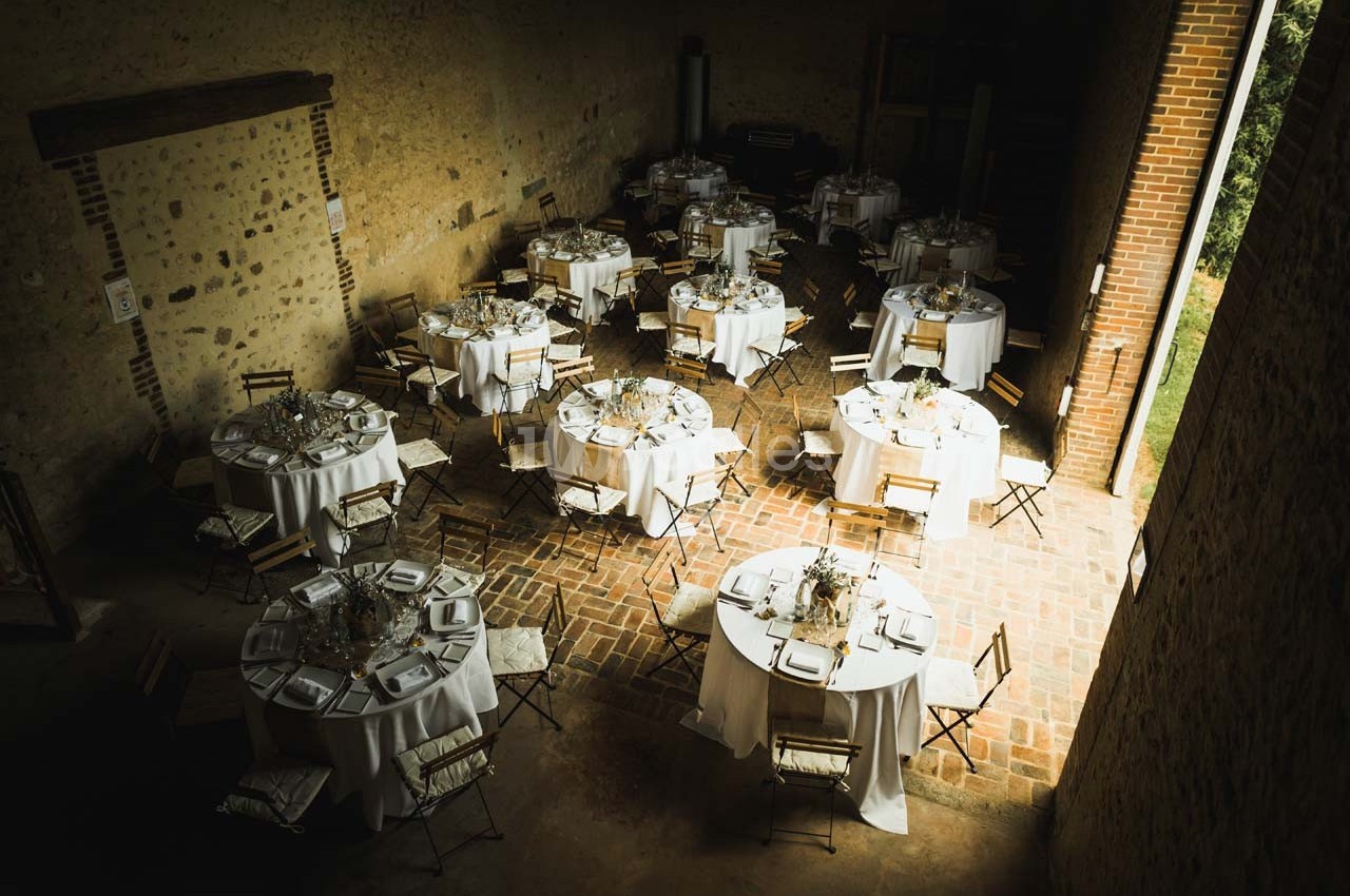 Tables rondes dressées avec nappes blanches et chaises en bois dans une salle en pierre au sol en briques.