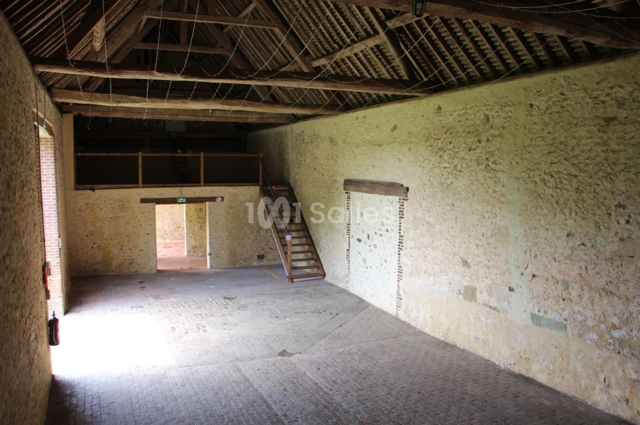 Intérieur d'une grange en pierre avec un sol en briques, un escalier en bois et une mezzanine.
