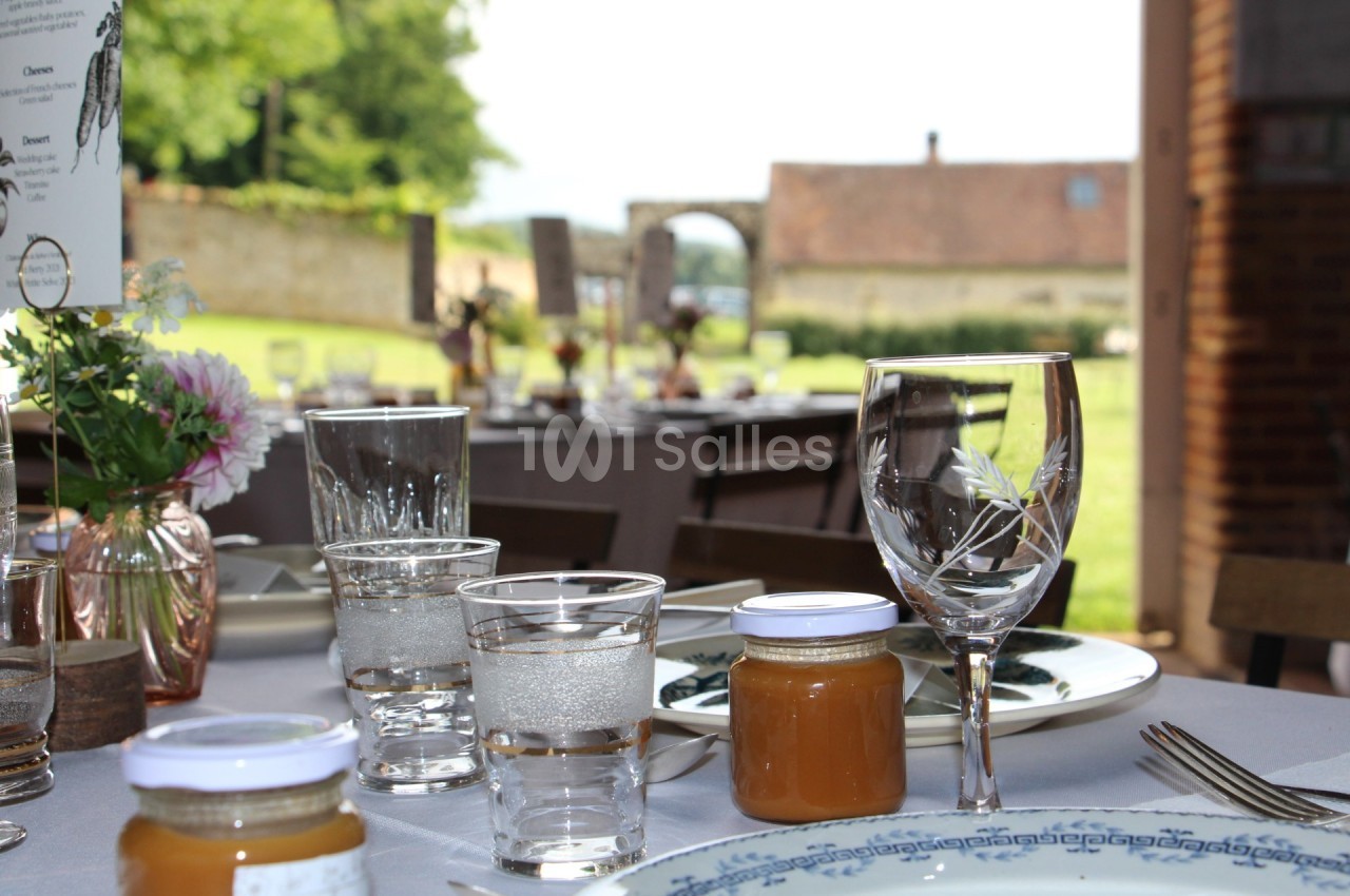 Table dressée avec vaisselle élégante, pots de confiture et verres, en extérieur avec vue sur un jardin et un bâtiment en…