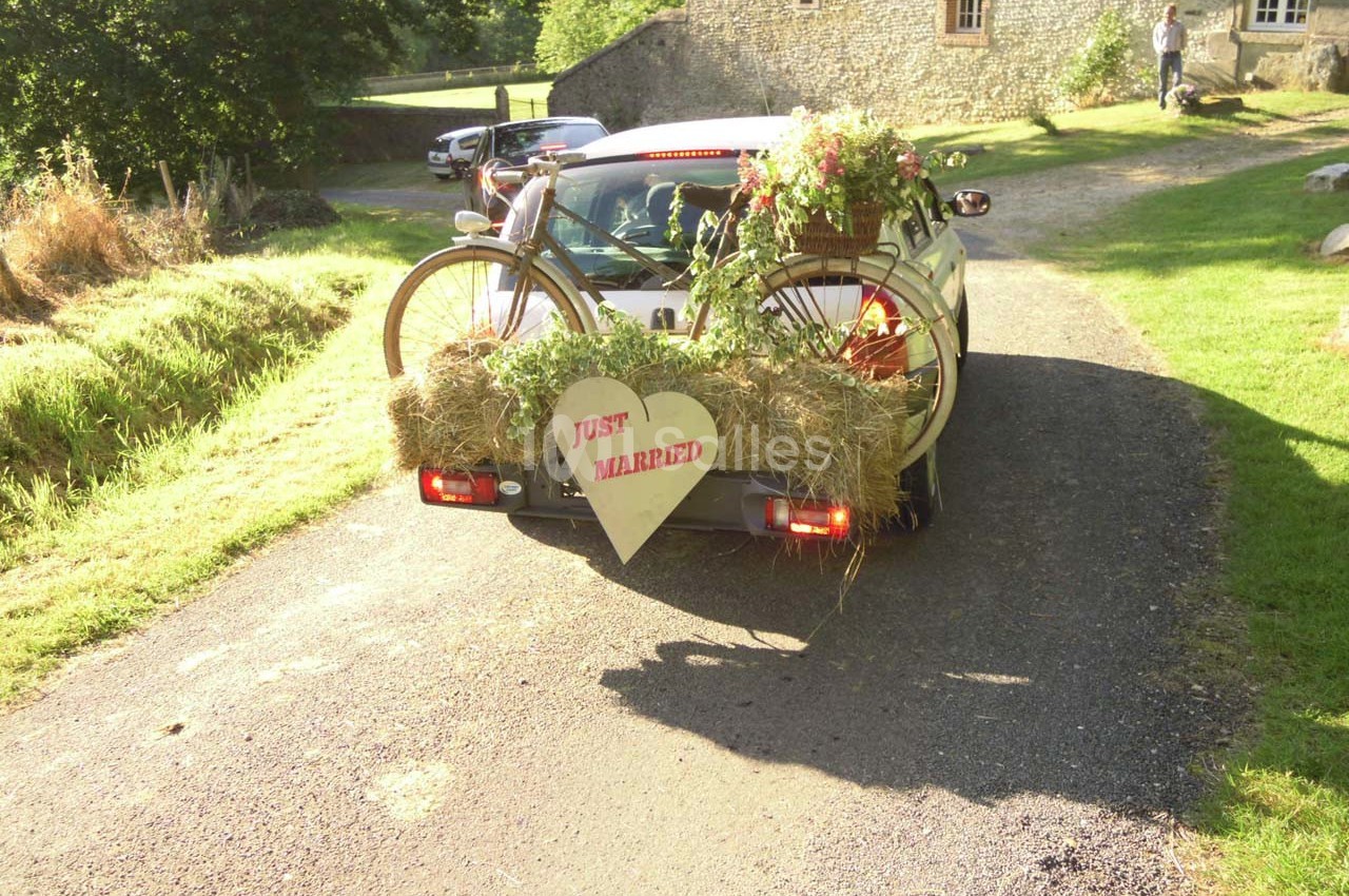 Voiture décorée avec des fleurs, du foin et un vélo, portant un panneau ’Just Married’, sur une route de campagne.
