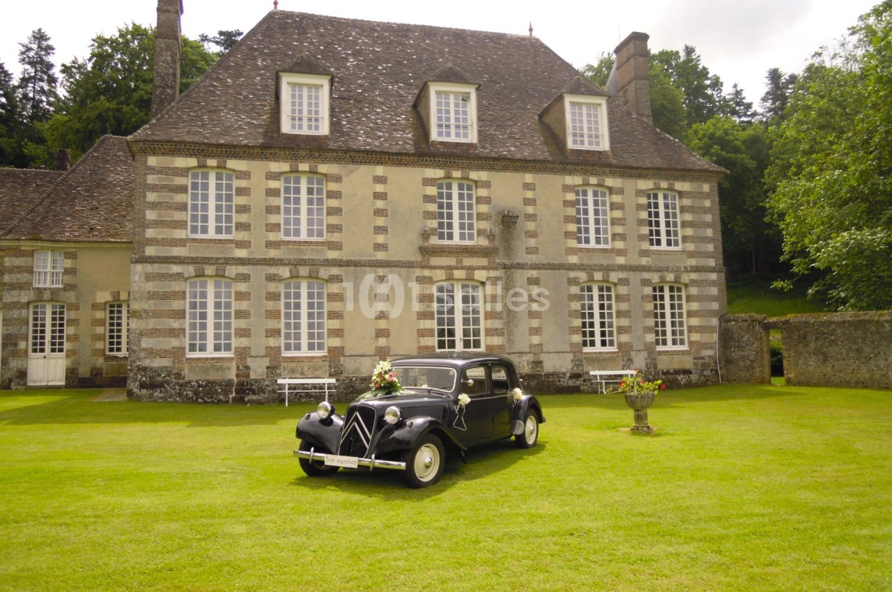 Voiture ancienne noire stationnée devant une grande maison en pierre avec jardin et arbres en arrière-plan.