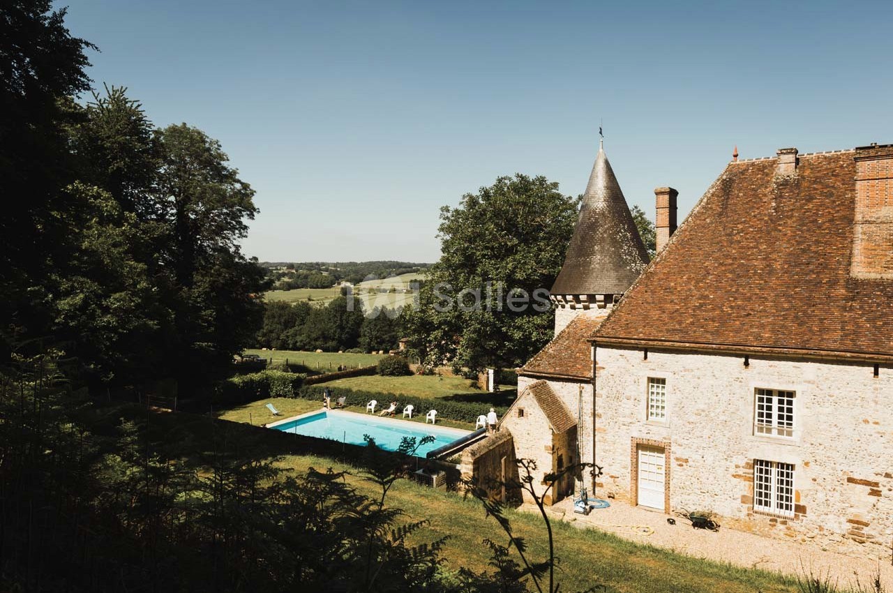Vue d’un bâtiment en pierre avec une tour, une piscine et un paysage champêtre en arrière-plan.