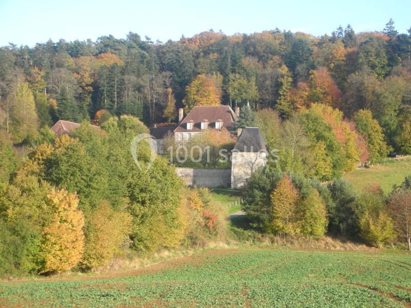 Manoir entouré d'arbres aux couleurs automnales, situé en lisière d'une forêt et bordé de champs.