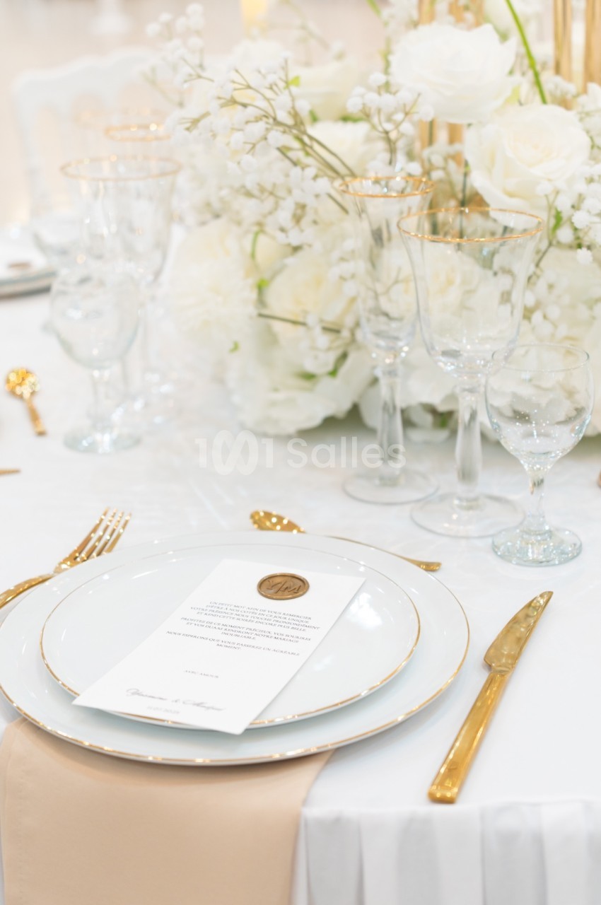 Table élégamment dressée avec vaisselle blanche et dorée, bouquet de fleurs blanches et menu posé sur une assiette.