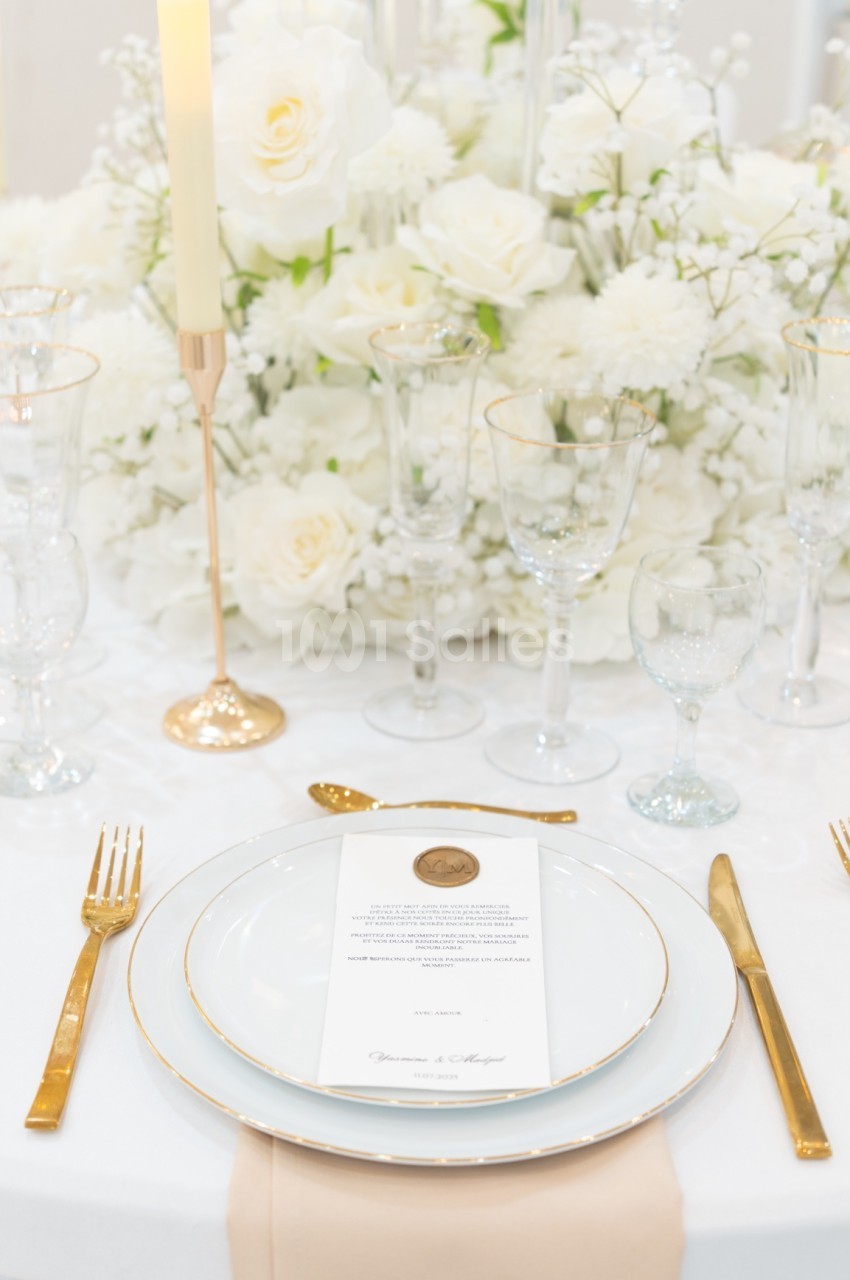 Table élégamment dressée avec vaisselle blanche et dorée, bougie allumée et centre de table floral blanc.