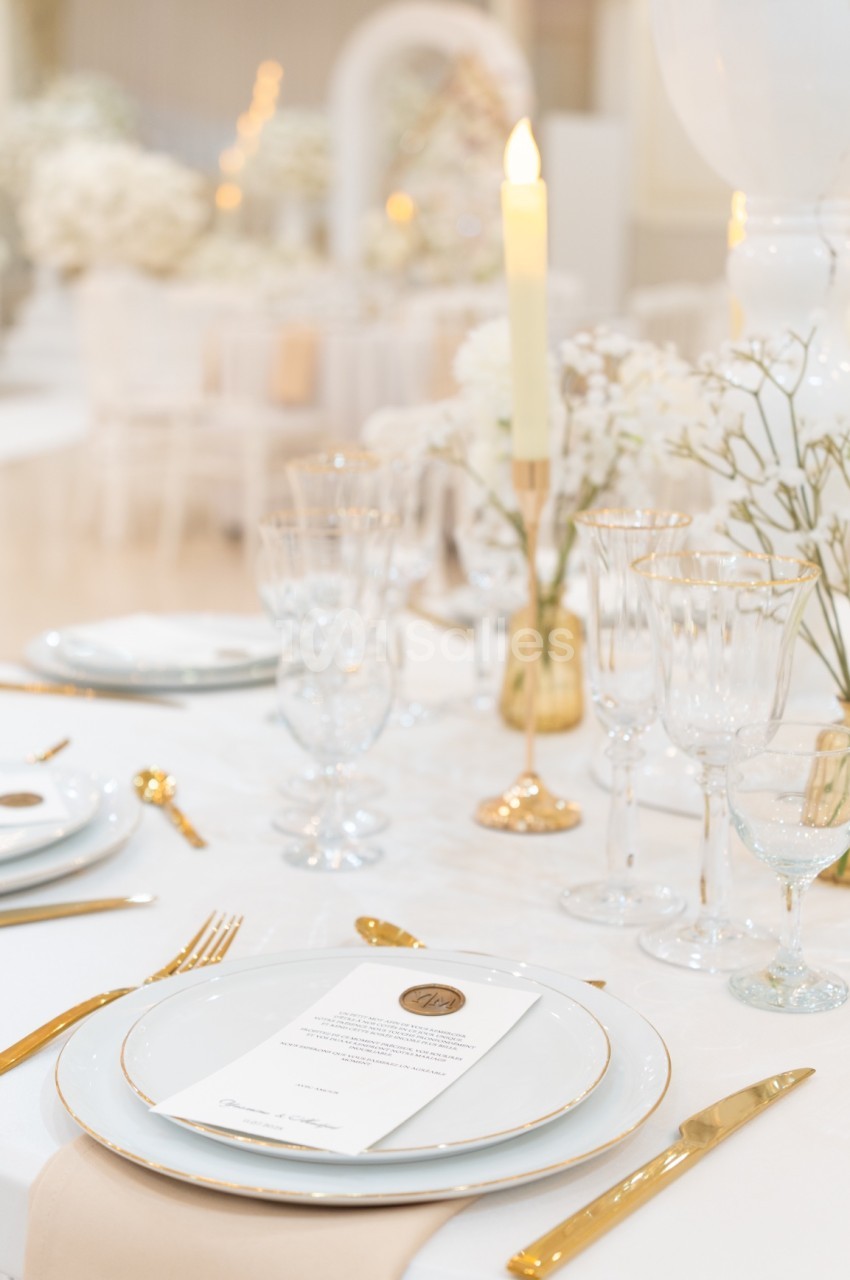 Table élégamment dressée avec vaisselle blanche, couverts dorés, bougie allumée et décorations florales blanches.