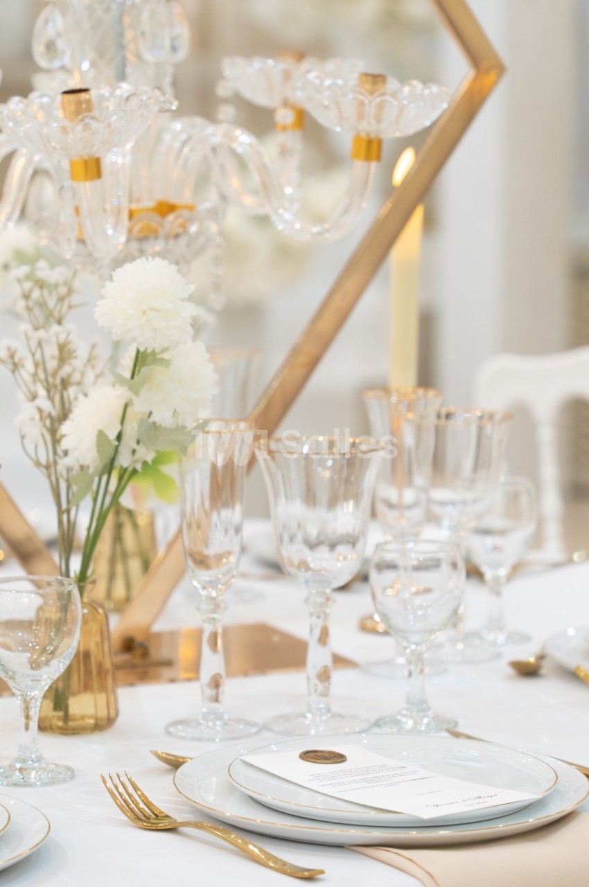 Table élégamment dressée avec vaisselle raffinée, chandelier en verre, fleurs blanches et décoration dorée.