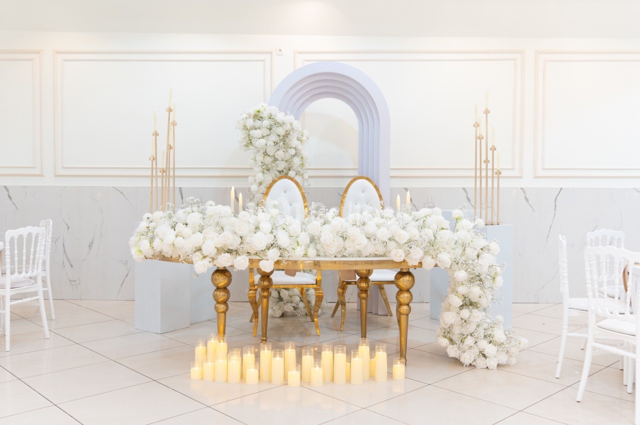 Table décorée de fleurs blanches, bougies au sol et arche florale en arrière-plan dans une salle lumineuse.
