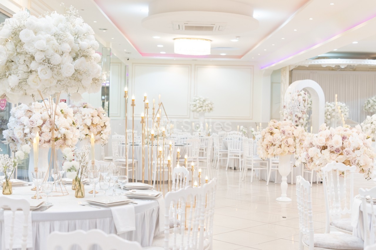 Salle de réception élégante décorée de fleurs blanches et roses, avec des tables dressées et des chandeliers dorés.