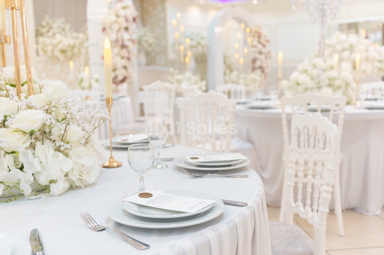 Salle de réception élégante décorée de fleurs blanches, tables dressées avec vaisselle raffinée et bougies allumées.