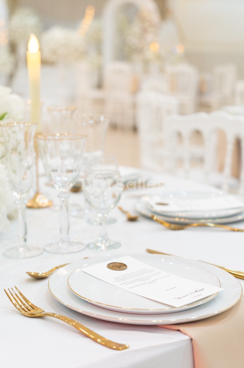 Table élégamment dressée avec assiettes, couverts dorés, verres en cristal et bougie allumée dans un décor lumineux.