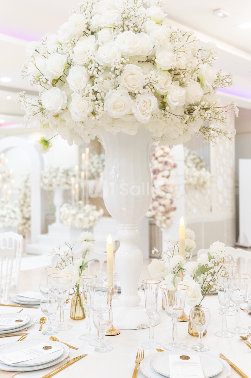 Centre de table avec un grand bouquet de roses blanches dans un vase, entouré de vaisselle élégante et de bougies.