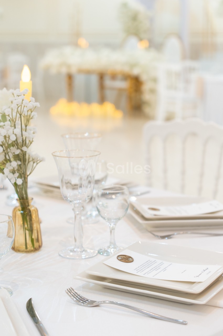 Table élégamment dressée avec vaisselle blanche, verres, menus et vase de fleurs blanches dans une salle lumineuse.