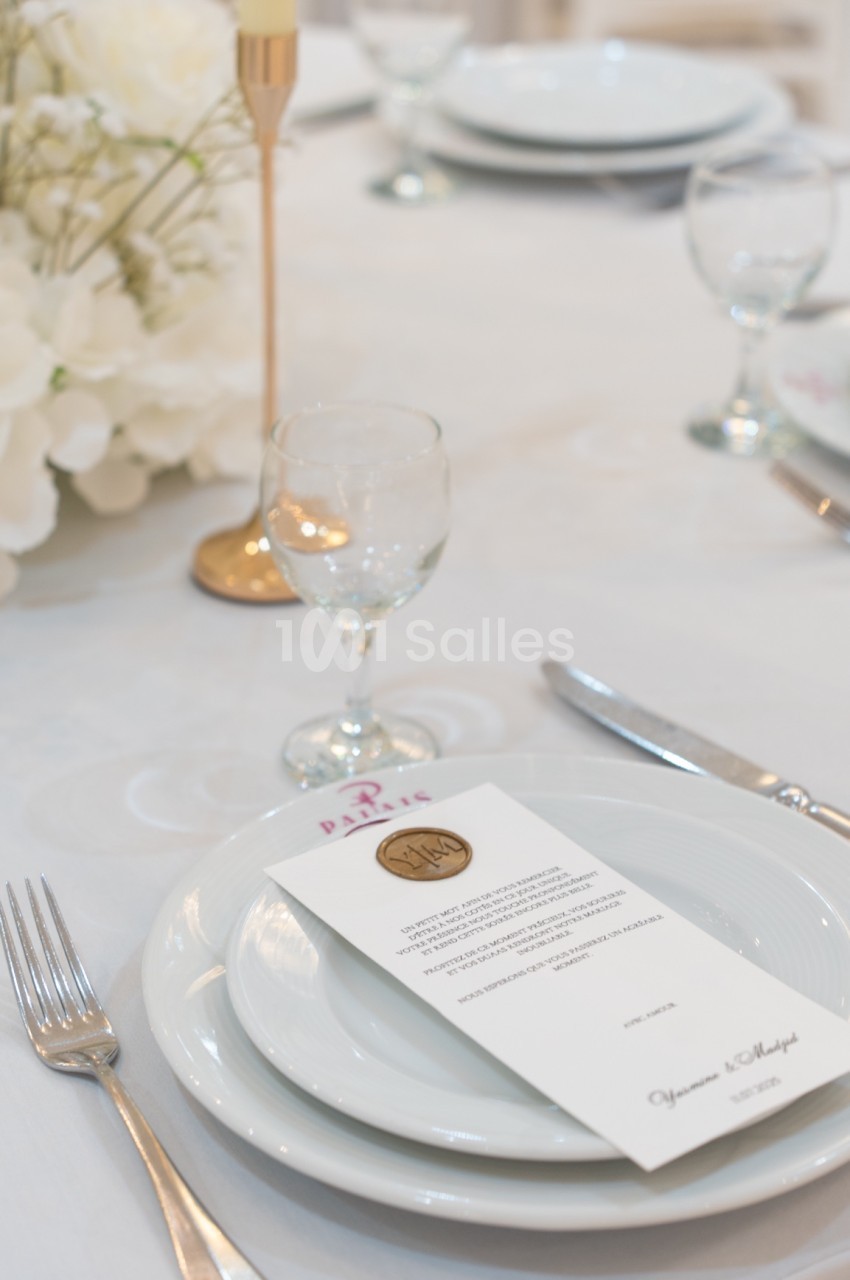 Table dressée avec assiette, couverts, verre, menu en papier et décoration florale blanche sur une nappe claire.