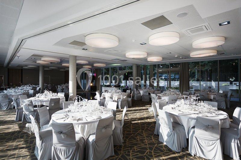 Salle de réception lumineuse avec tables rondes dressées, nappes blanches et chaises couvertes, prête pour un événement.