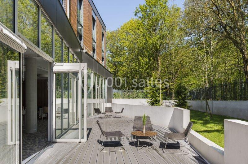 Terrasse en bois avec chaises et table, bordée par un bâtiment moderne et un jardin arboré en arrière-plan.