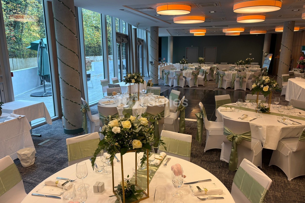 Salle de réception décorée pour un événement, avec tables rondes dressées, nappes blanches et compositions florales.