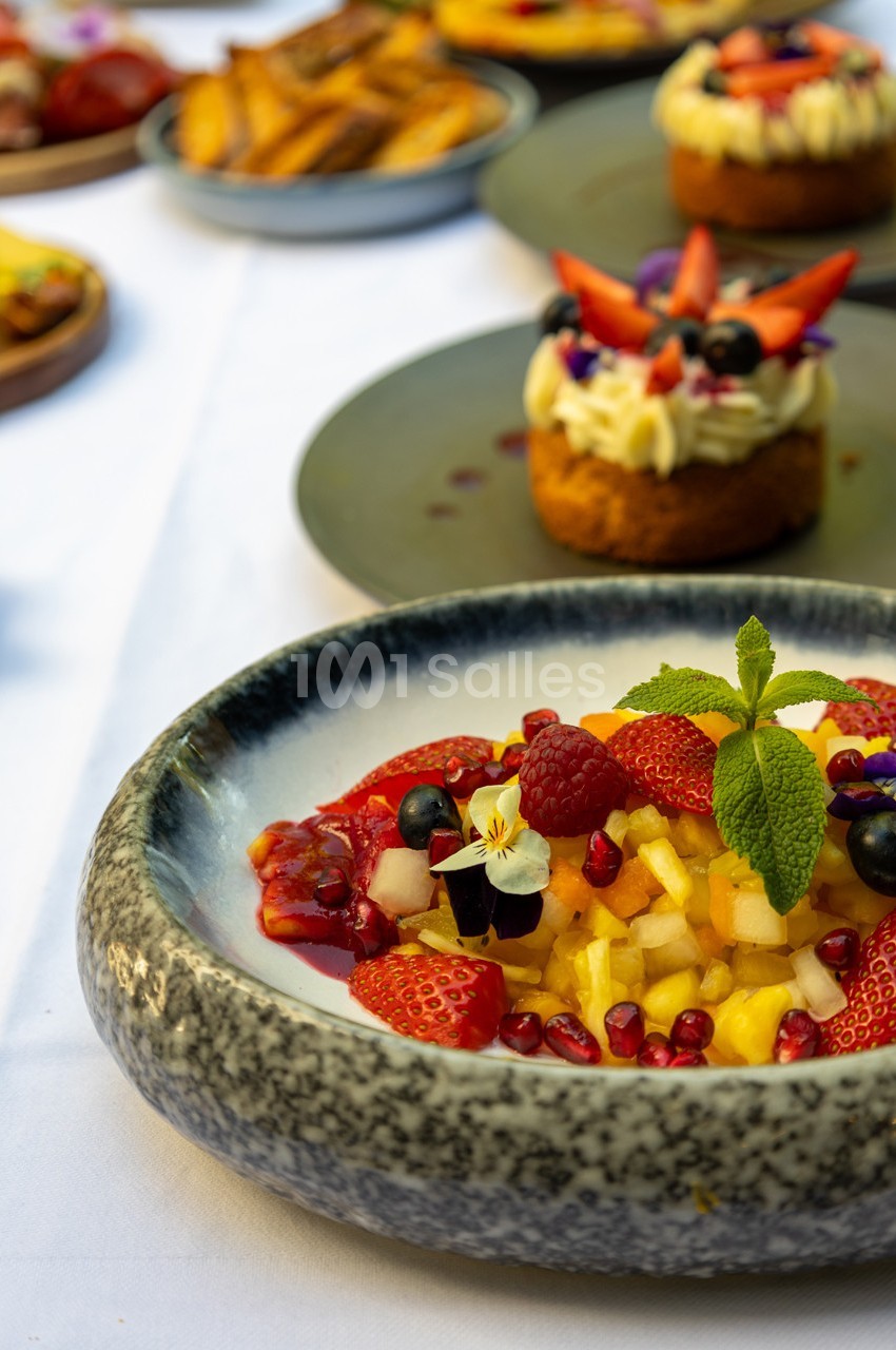 Salade de fruits frais garnie de menthe, avec des desserts et des frites en arrière-plan sur une table dressée.