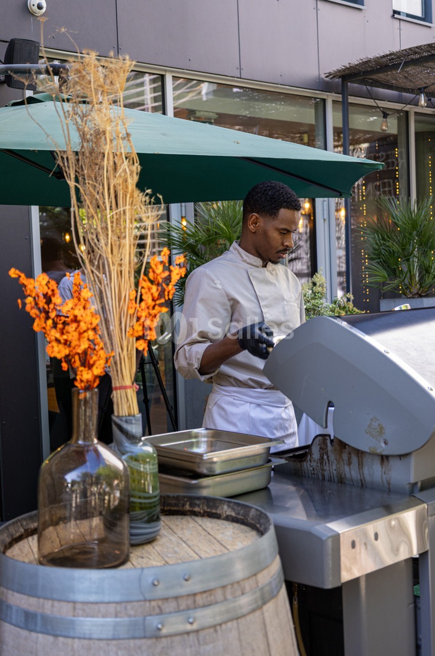 Un chef cuisine sur un barbecue en extérieur, près d'une décoration avec des fleurs séchées et des plantes.