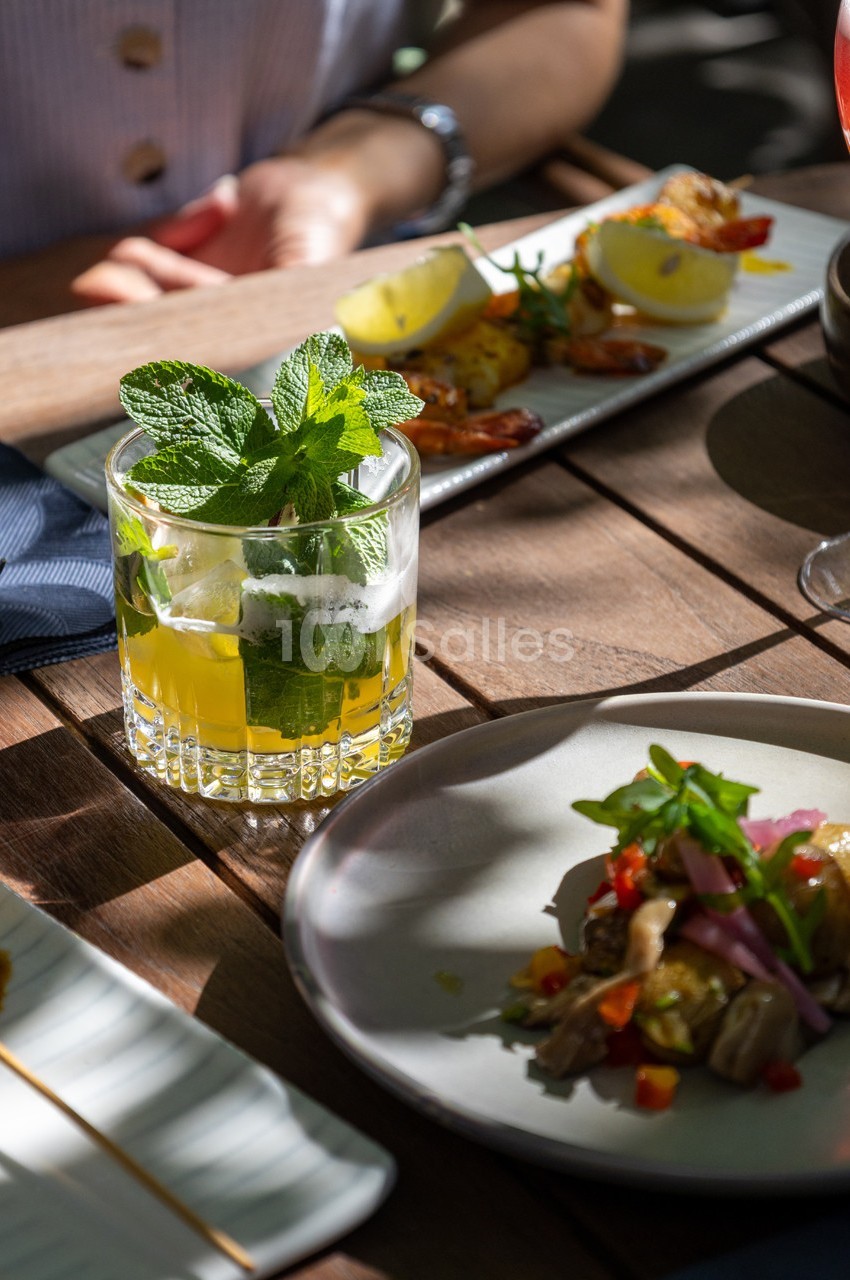 Verre de cocktail à la menthe sur une table en bois, accompagné de plats variés et éclairé par une lumière naturelle.