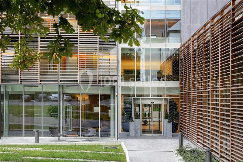 Façade moderne d'un bâtiment avec grandes baies vitrées, brise-soleil en bois et espace extérieur aménagé.