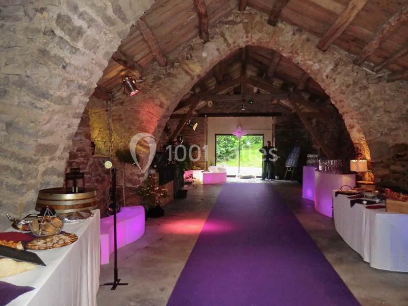 Salle voûtée en pierre avec tables de buffet éclairées, tapis violet au centre et vue sur un jardin à l'arrière.