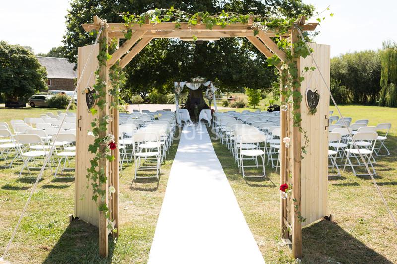 Allée centrale décorée pour une cérémonie en plein air, bordée de chaises blanches et d'une arche en bois ornée de feuillage.