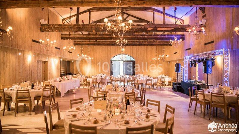 Salle de réception décorée avec des tables rondes dressées, éclairage chaleureux et poutres apparentes au plafond.