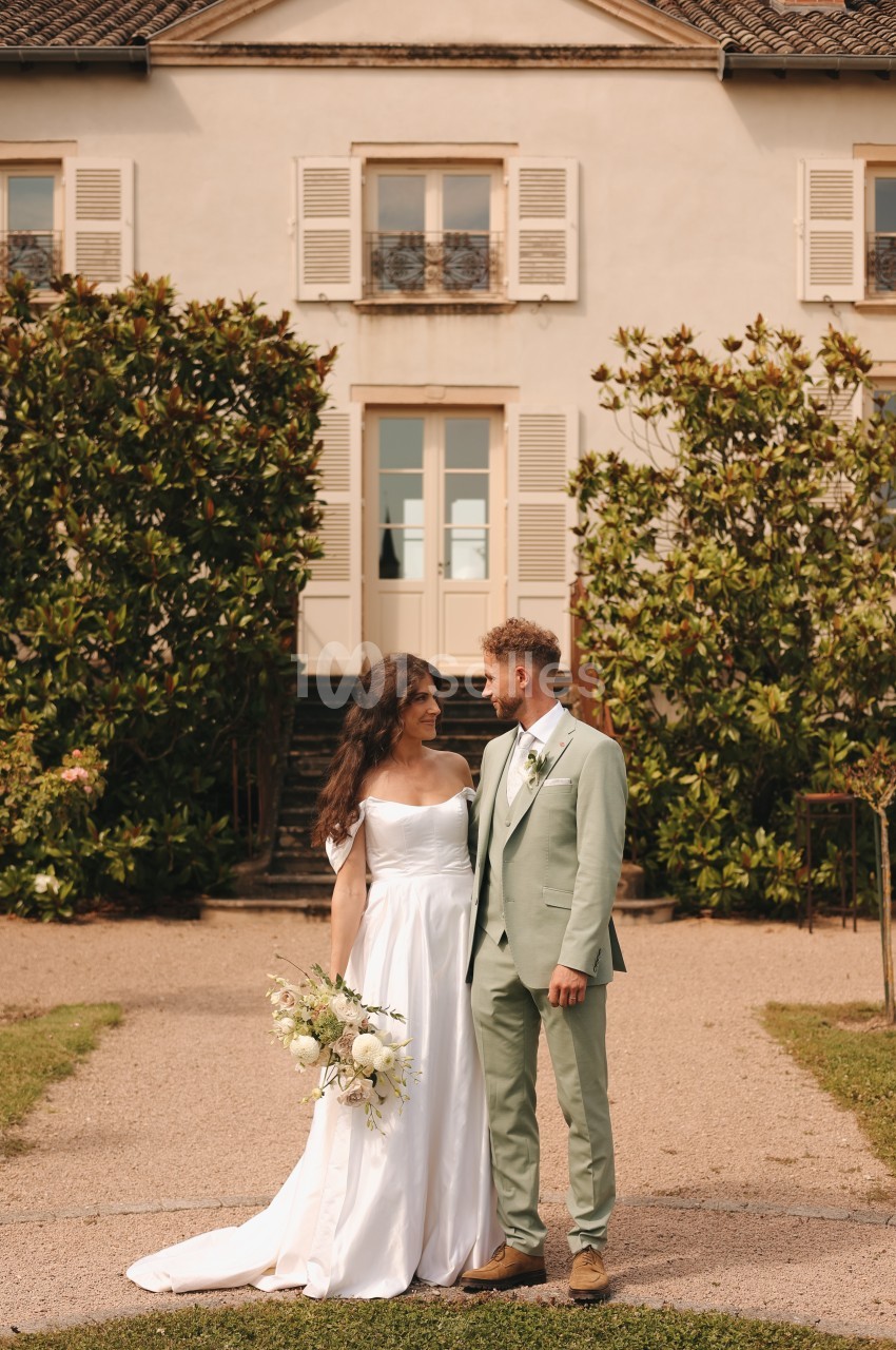 Un couple en tenue de mariage se tient devant une maison entourée de verdure, se regardant avec complicité.