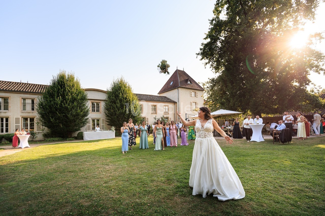 Une mariée lance son bouquet devant des invitées rassemblées dans le jardin d'un domaine sous un soleil couchant.