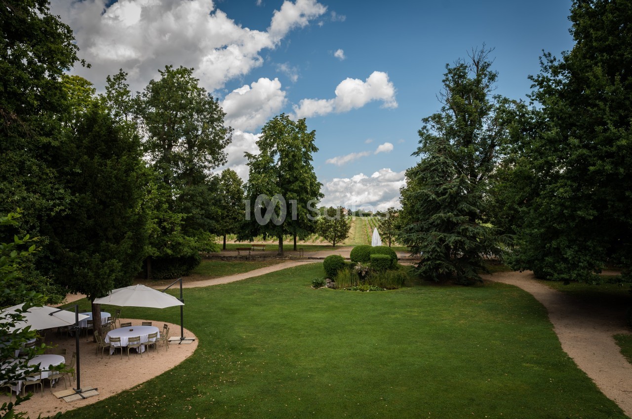 Jardin verdoyant avec pelouse, arbres, chemins en gravier et tables sous parasols par une journée ensoleillée.