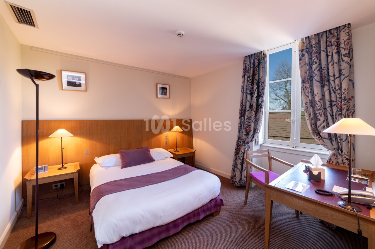 Chambre d'hôtel lumineuse avec lit double, bureau en bois, lampes allumées et fenêtre donnant sur un extérieur arboré.