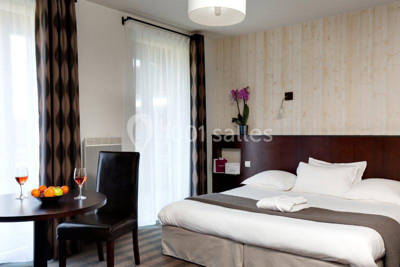 Chambre d'hôtel avec lit double, table ronde, chaise, orchidée en décoration et lumière naturelle provenant des fenêtres.